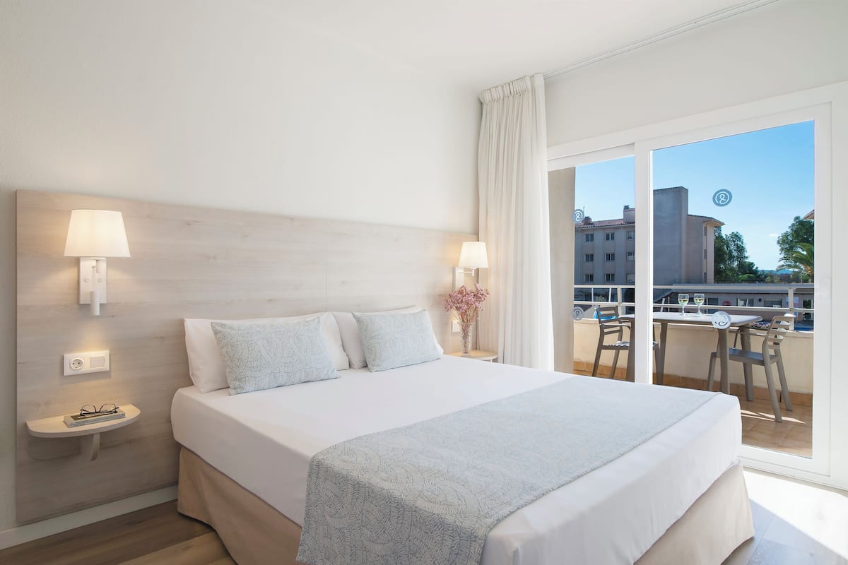 Een hotelkamer met een groot bed, twee bedlampen, een nachtkastje en uitzicht op een balkon met zitplaatsen.