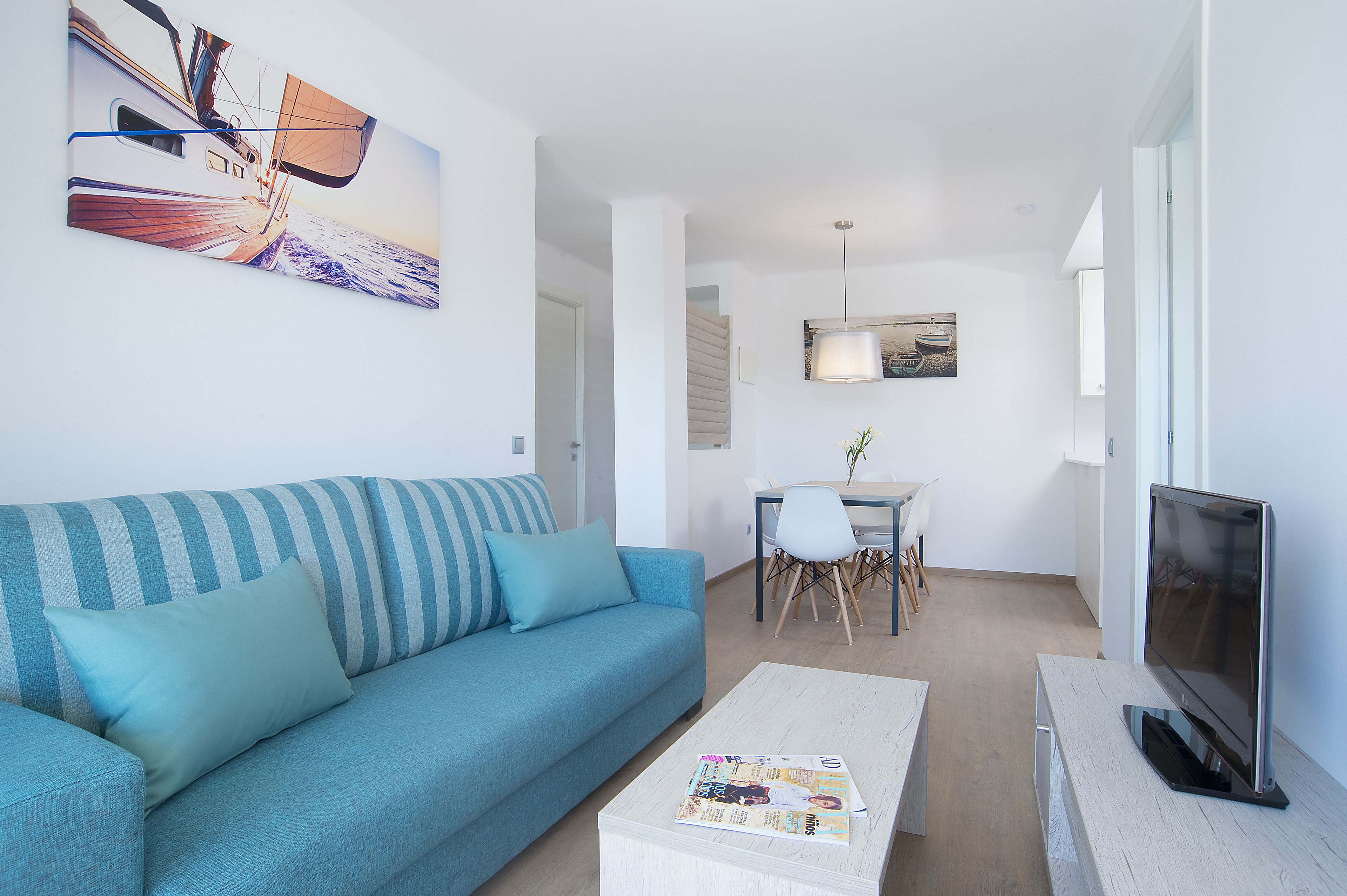 A modern living room with a blue sofa, a coffee table with a magazine, a flat-screen TV, and a dining area with a table and chairs.