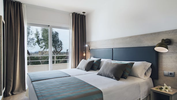 Une chambre d’hôtel avec un grand lit, deux tables de chevet, une commode, une fenêtre avec des rideaux et une vue sur les arbres à l’extérieur.
