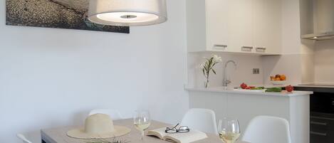 A modern dining area with a table set for two, white chairs, a hanging light fixture, and a kitchen in the background.