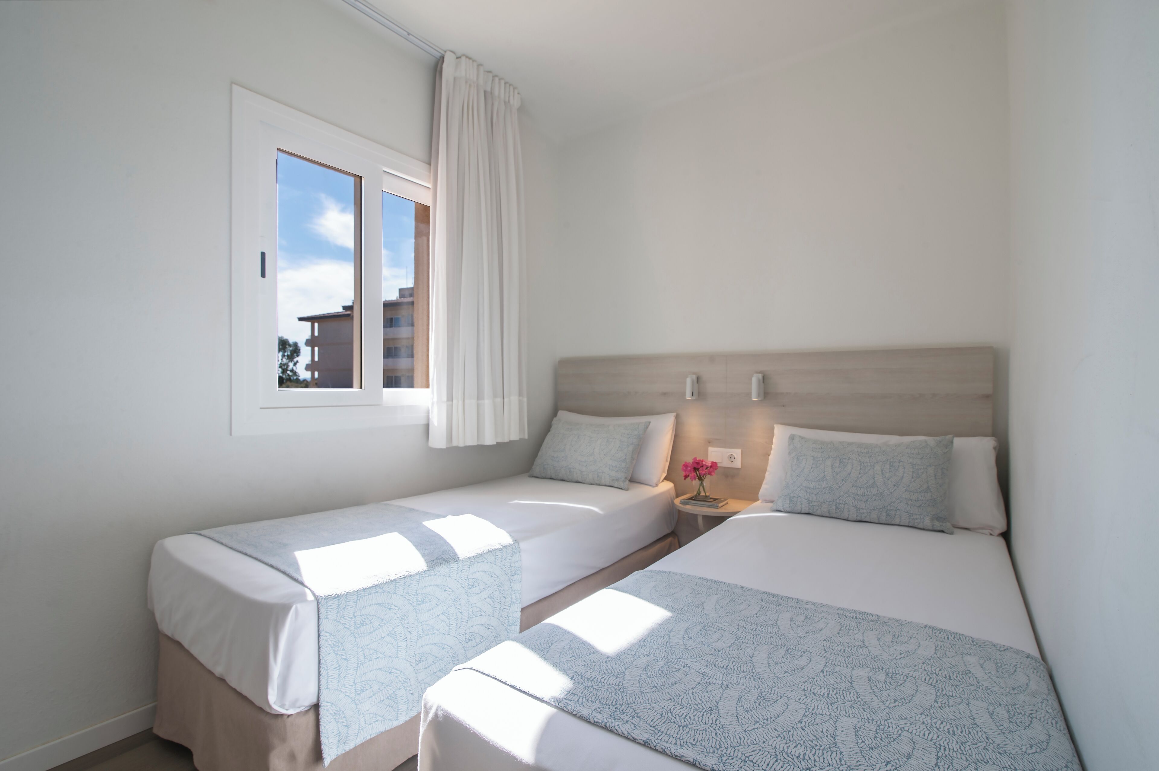 Habitación de hotel con dos camas individuales, una mesita de noche con un jarrón de flores, una ventana con cortinas y vistas a edificios.