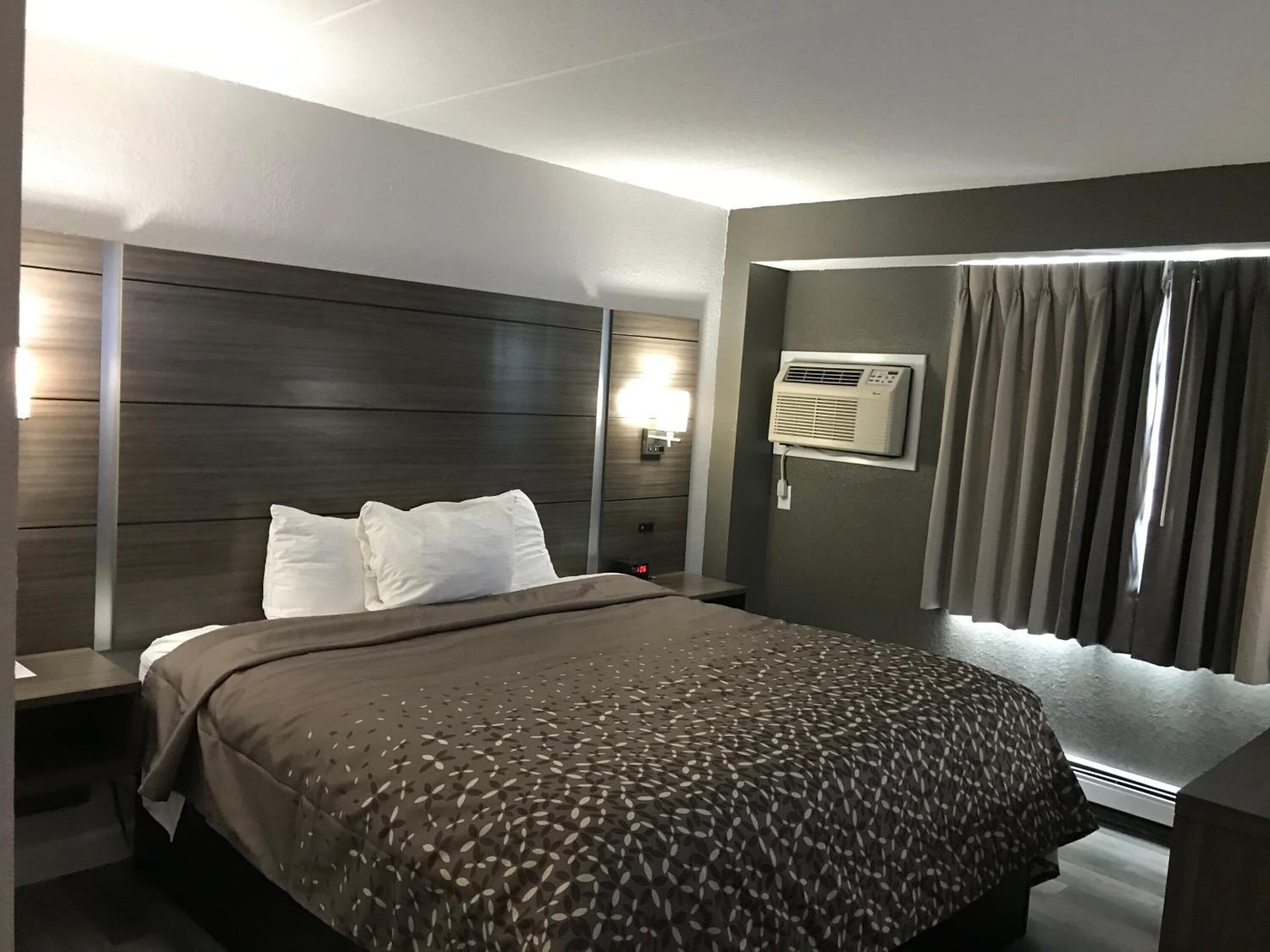 Una habitación de hotel moderna con una cama grande, una mesita de noche, aire acondicionado de pared y una ventana con cortinas.