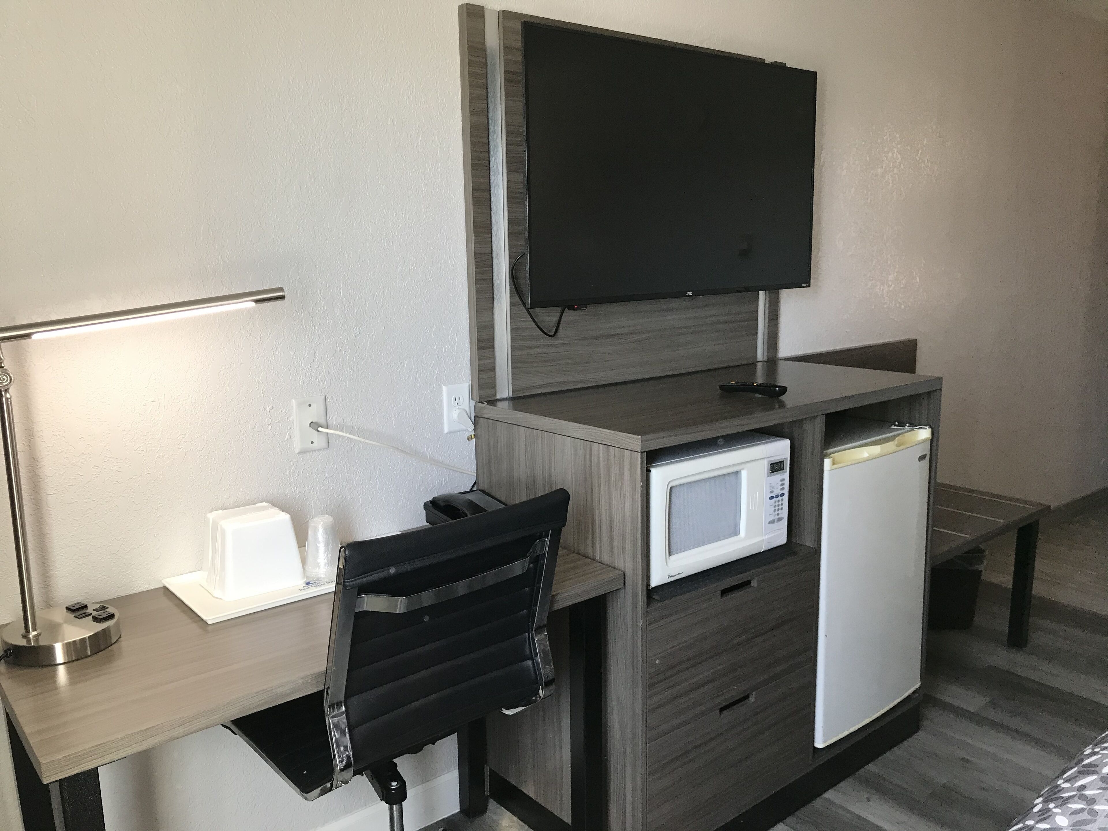 A hotel room with a desk, a chair, a television mounted on the wall, a microwave, and a mini-fridge.