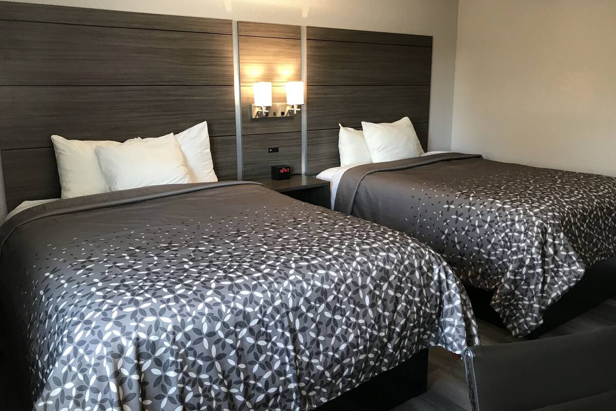 A hotel room with two beds, dark wood headboard, nightstands with lamps, and a digital clock.