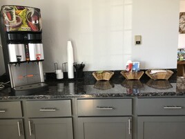Una zona de cocina con cafetera, vasos de papel y dispensadores de condimentos.