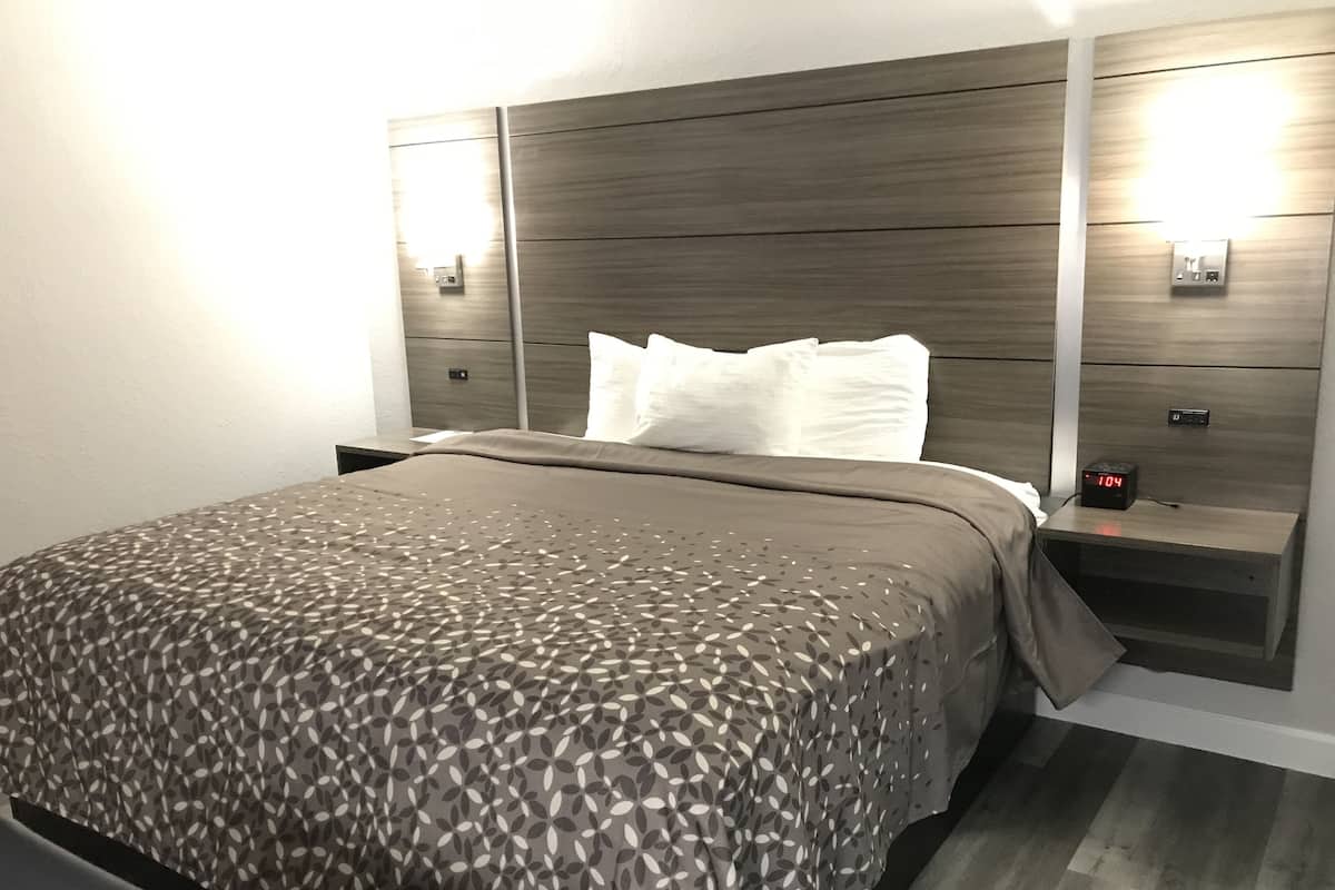 A modern hotel room with a large bed, a nightstand with a clock, and wall-mounted lighting.