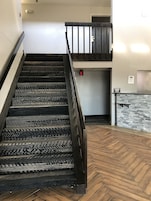 Una escalera con barandal de madera y alfombra estampada, que conduce a una habitación con pared de piedra y un letrero que indica una máquina de hielo.