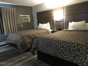 A hotel room with two beds, a headboard with a mirror, an air conditioning unit, and a bedside table with a clock.