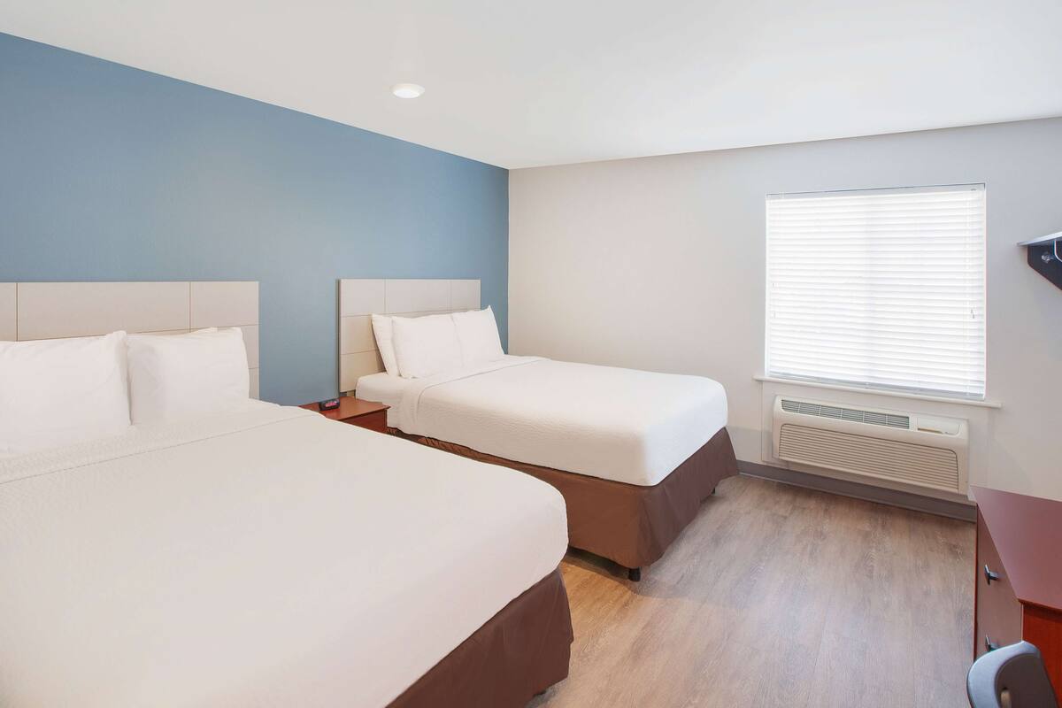 A hotel room with two beds, a wooden floor, a window with blinds, and a built-in heater.