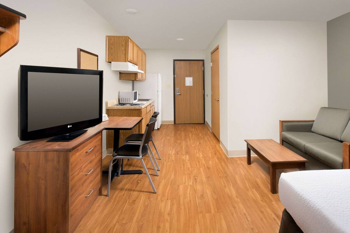 A compact hotel room with a wooden floor, a TV on a wooden stand, a small kitchenette, a sofa, and a bed.