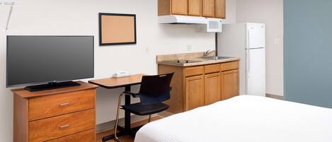 Habitación de hotel pequeña y funcional con cama, escritorio y silla, televisor, kitchenette con microondas y refrigerador, y armarios de madera.