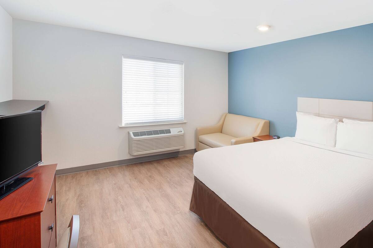 A hotel room with a bed, a TV, a chair, and a window with blinds.