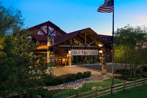 Una cabaña con una destacada bandera estadounidense, una entrada amplia con un letrero que dice "Great Wolf Lodge" y un jardín delantero bien cuidado con una valla de madera.