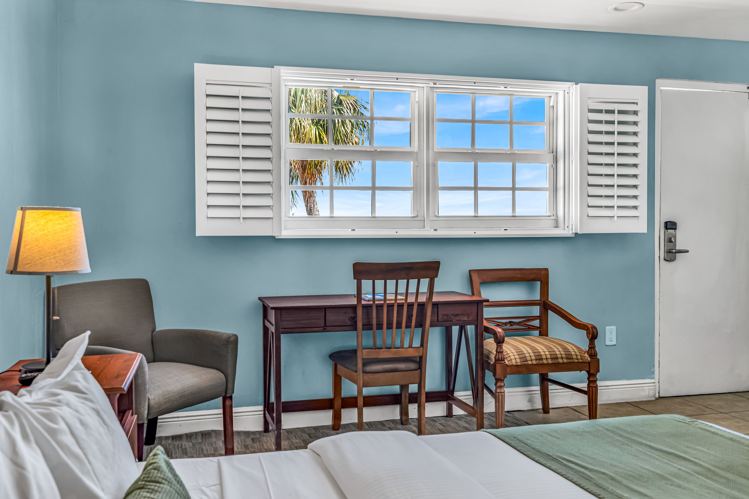 Una habitación con una cama, un escritorio, dos sillas, una lámpara y una ventana con contraventanas.