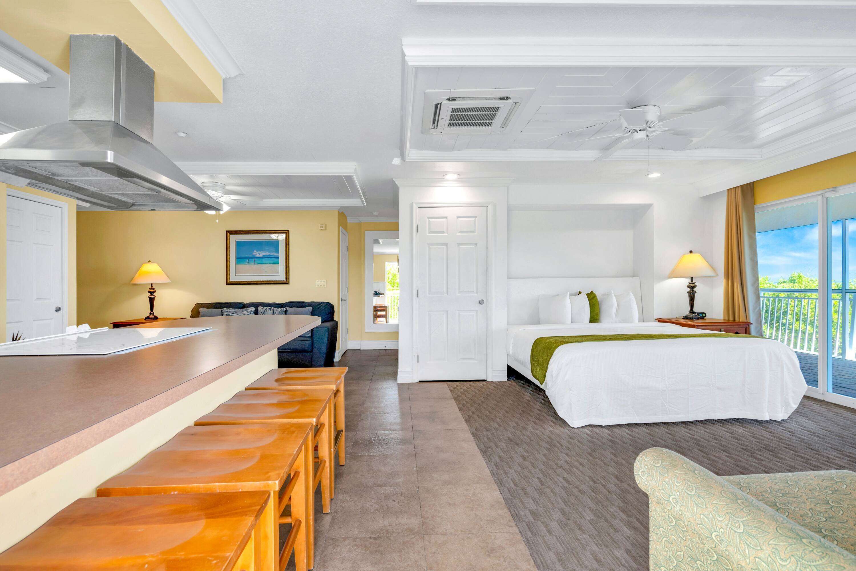 A hotel room with a kitchenette, a bed with a green throw, a balcony with a view, and a dining area with wooden stools.