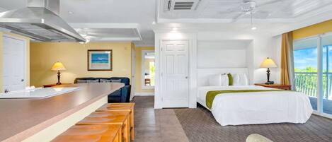 Habitación de hotel con kitchenette, cama con manta verde, balcón con vistas y zona de comedor con taburetes de madera.