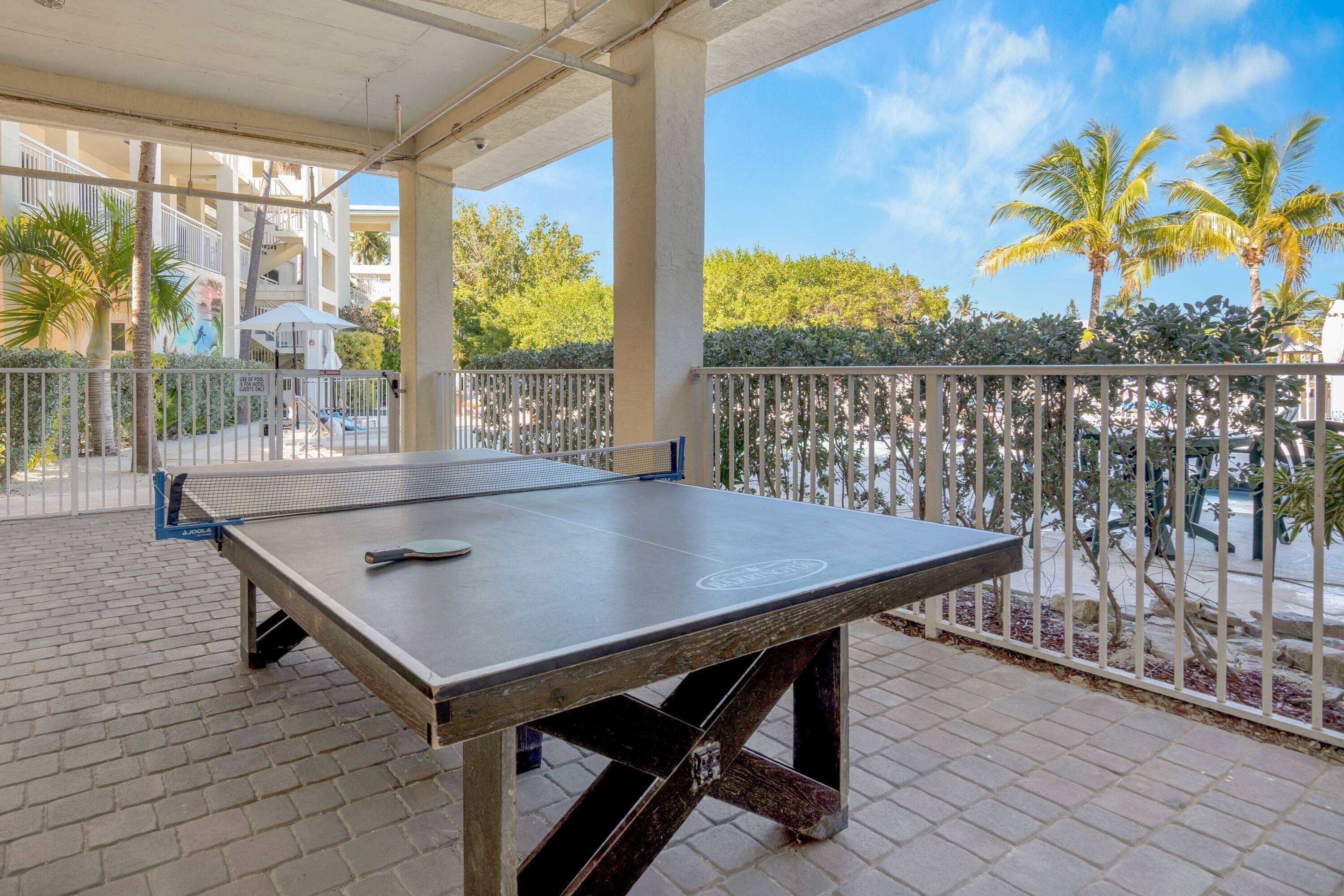 A ping pong table on a covered patio with a view of palm trees and a pool.