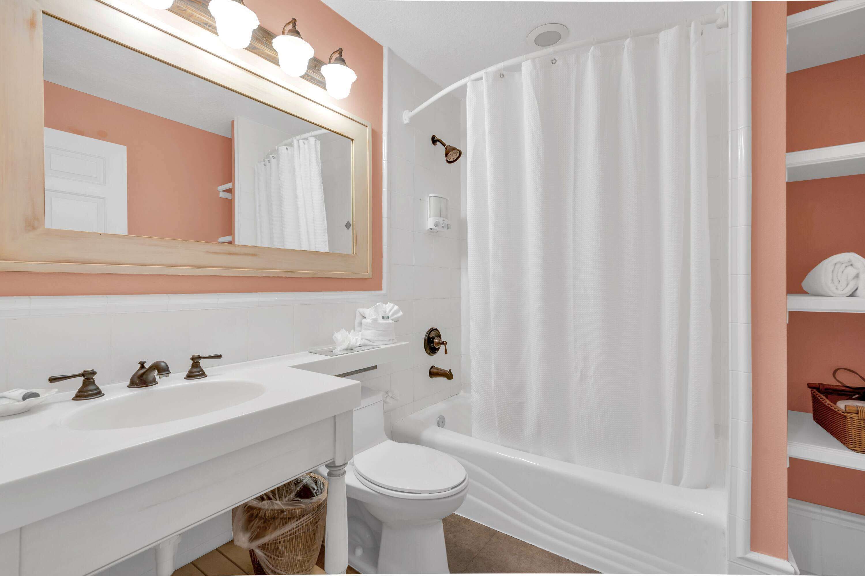A bathroom with a white sink, a toilet, a bathtub with a shower curtain, a mirror with lights, and a shelf with towels.