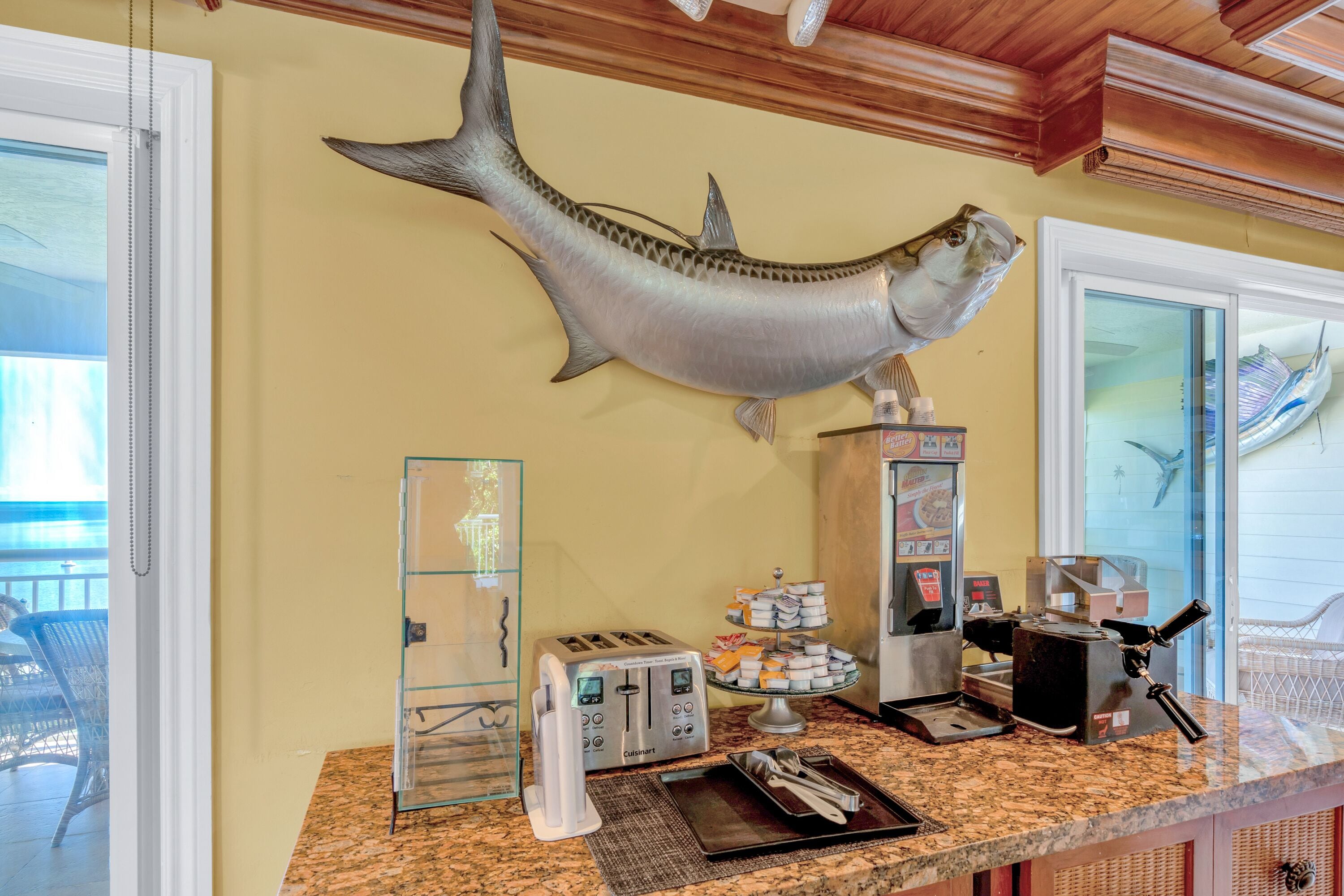 A kitchen counter with a toaster, a coffee maker, a tray, and a large fish mounted on the wall.