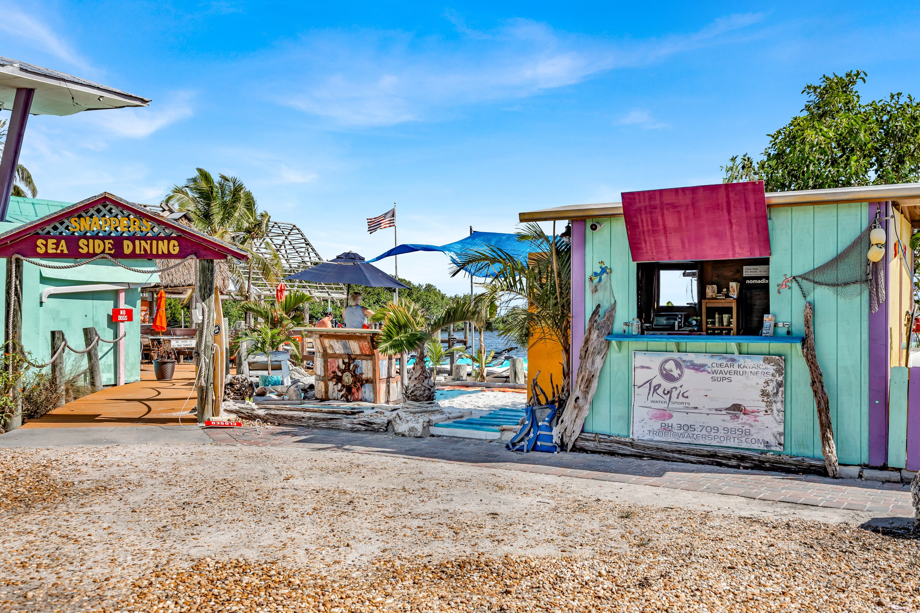 Zona de comedor junto a la playa, con un letrero que dice 'Sea Side Dining' y un cartel de 'Tropical Sports'.