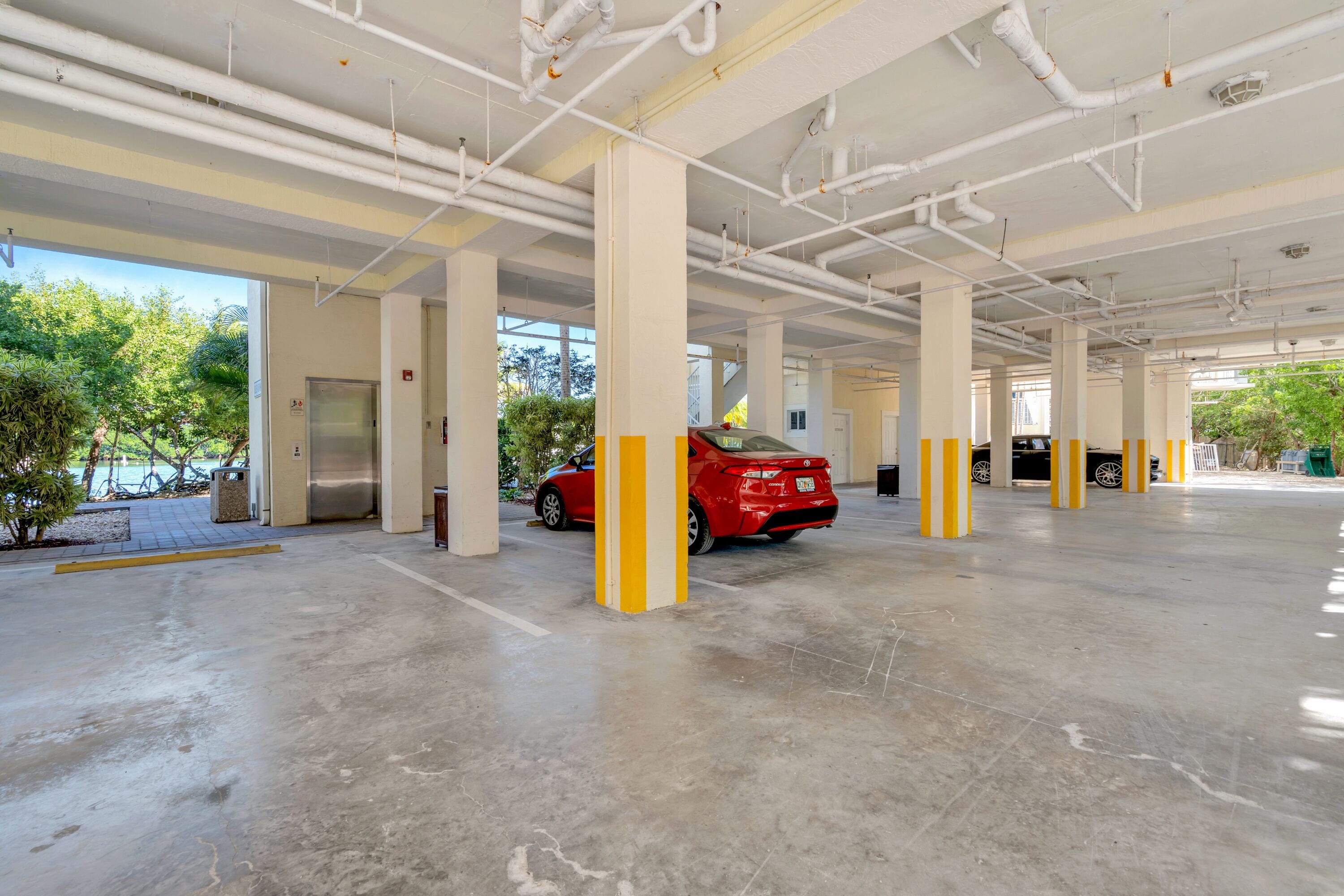 A spacious parking garage with multiple columns, a red car parked, and a clear view of trees outside.