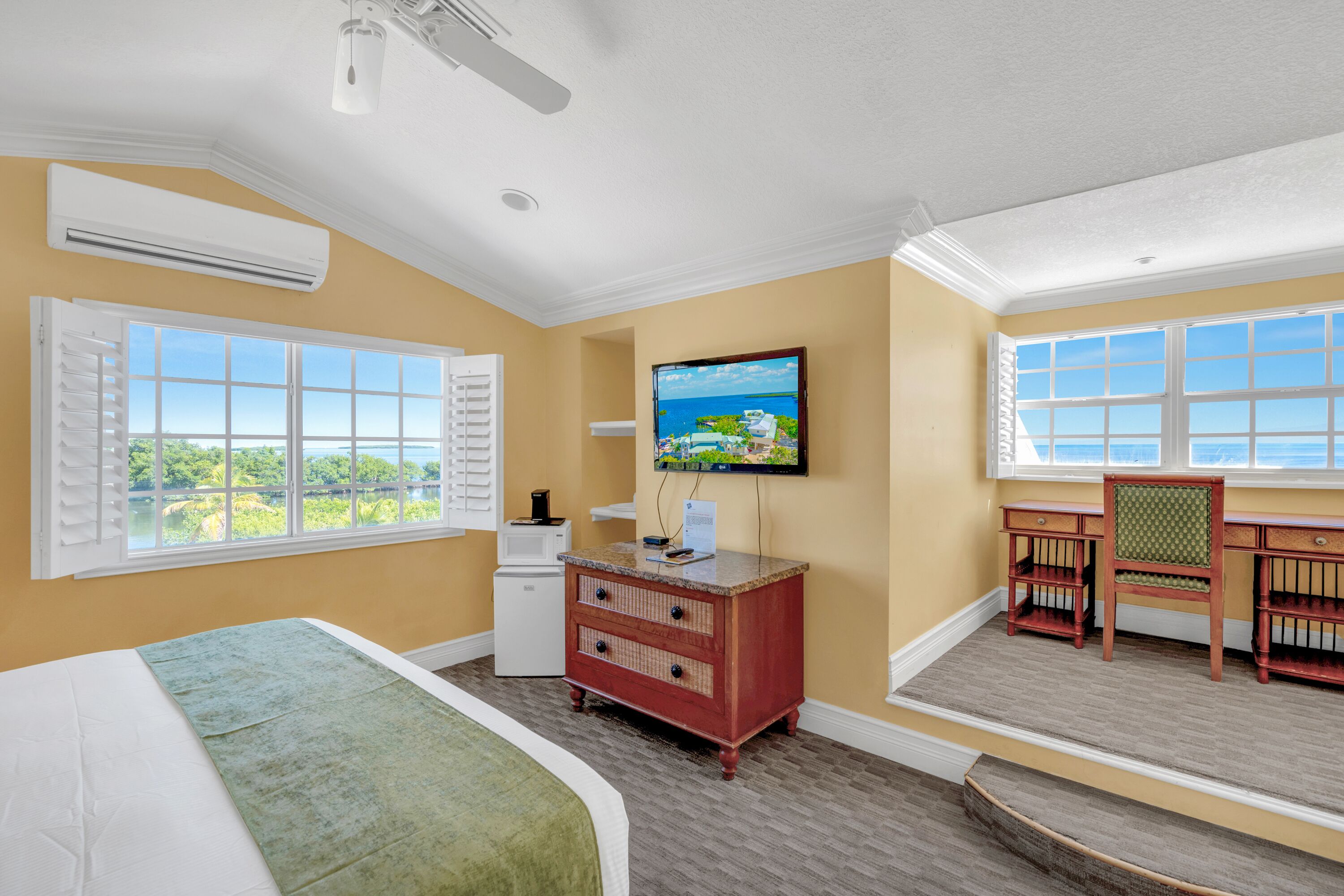 Habitación de hotel con cama, escritorio, silla, cómoda, televisor y ventana con vista a vegetación.