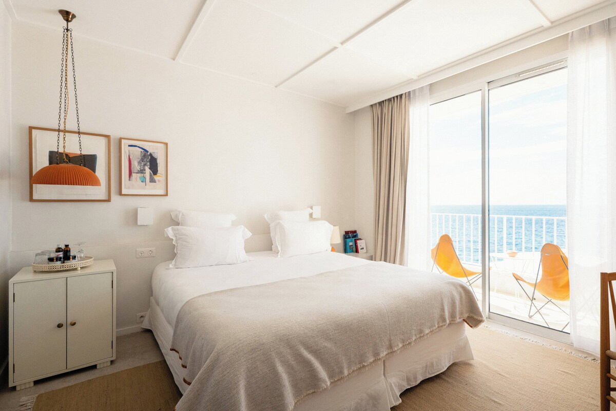A hotel room with a large bed, a nightstand, a small cabinet, a window with a view of the sea, and a chair by the window.