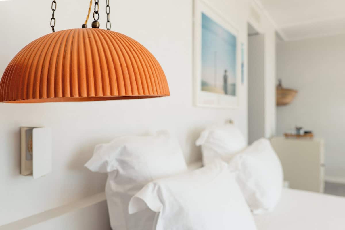 Quarto com cama branca, almofadas brancas e luminária pendente laranja.