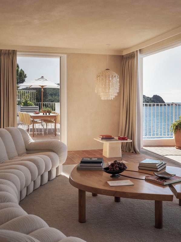 Una sala de estar moderna con un sofá, una mesa de centro redonda de madera y vista al mar a través de grandes ventanales.