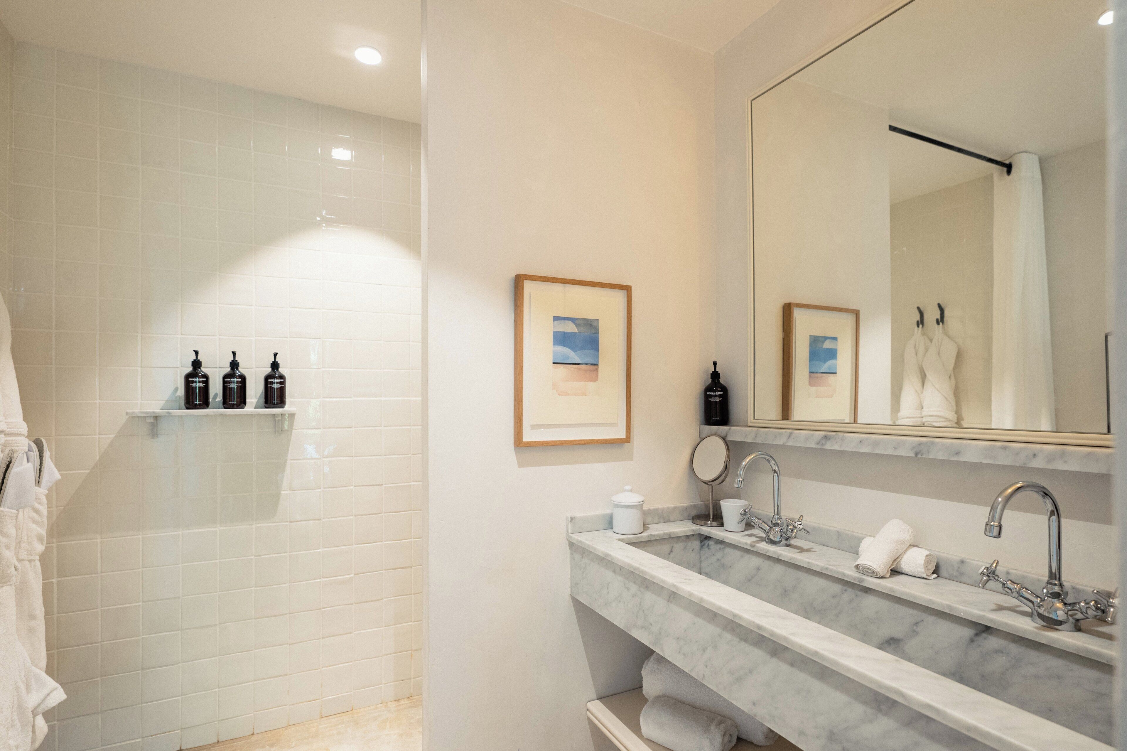 A modern bathroom with white tiles, a large mirror, two sinks, and a shower area.
