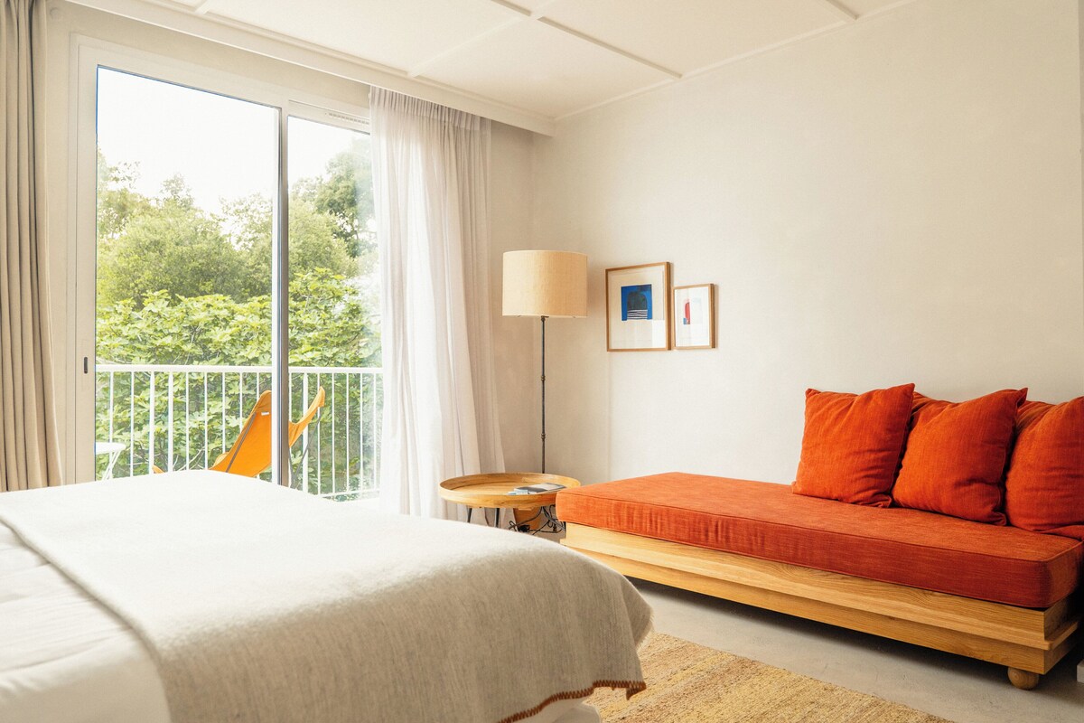 A hotel room with a bed, a sofa, a balcony with a chair, and a small table with a lamp.