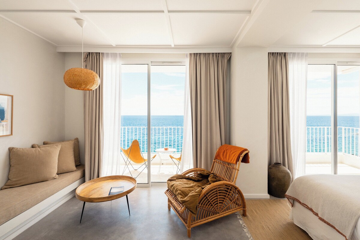 A hotel room with a balcony, a wicker chair, a sofa, and a bed.