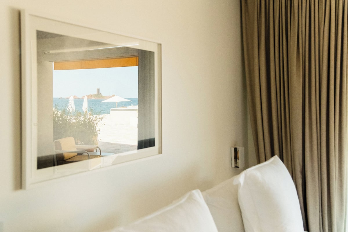 Quarto de hotel com quadro de uma paisagem costeira, cama com lençóis brancos e cortina na janela.