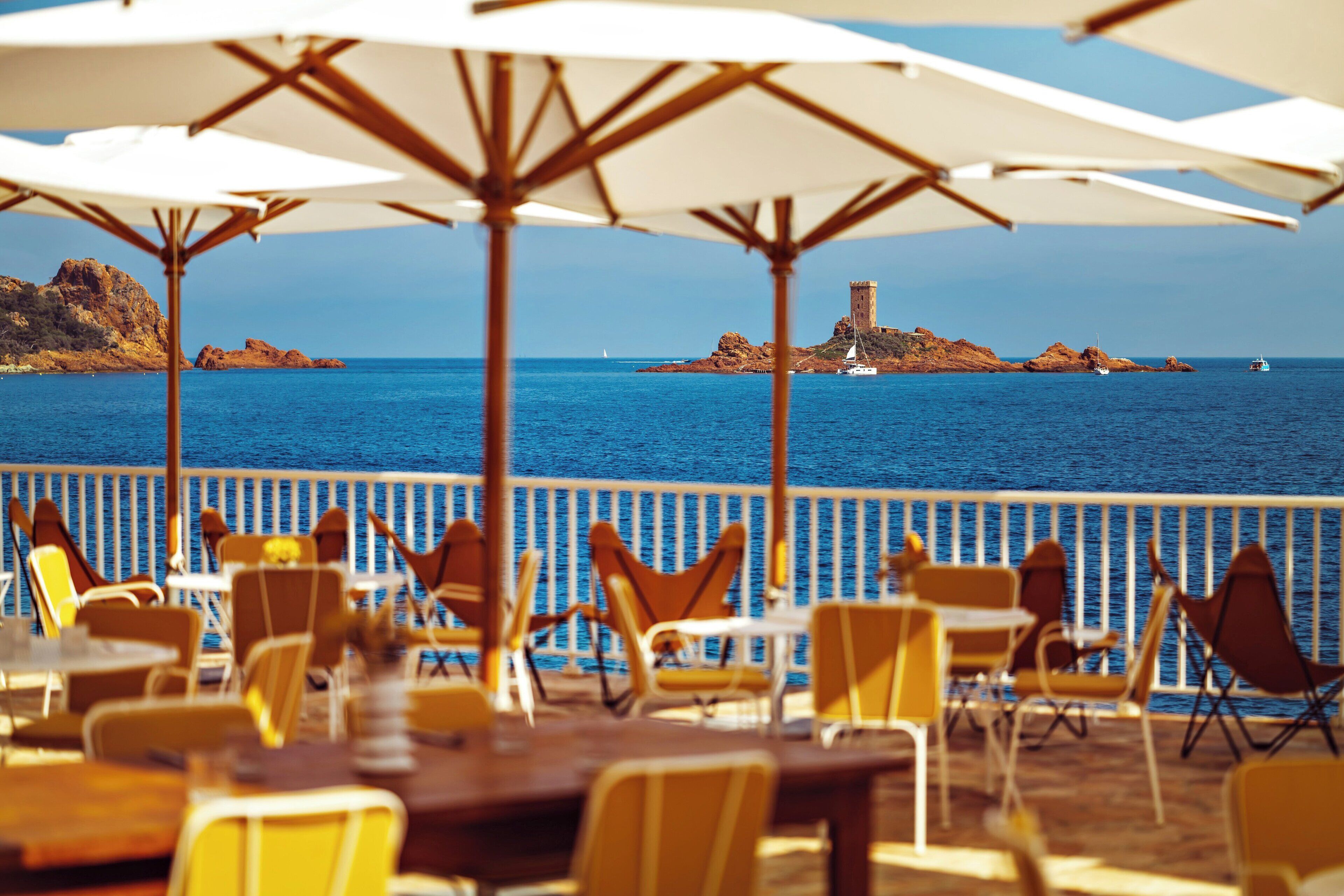 Een restaurant aan de kust met terras, parasols en uitzicht op zee en rotsformaties.