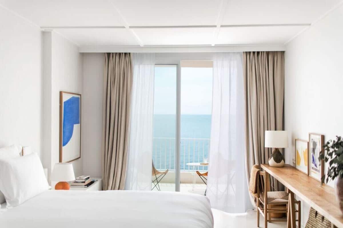 Quarto de hotel com uma cama, uma escrivaninha com abajur, uma cadeira e vista para o mar por grandes janelas.