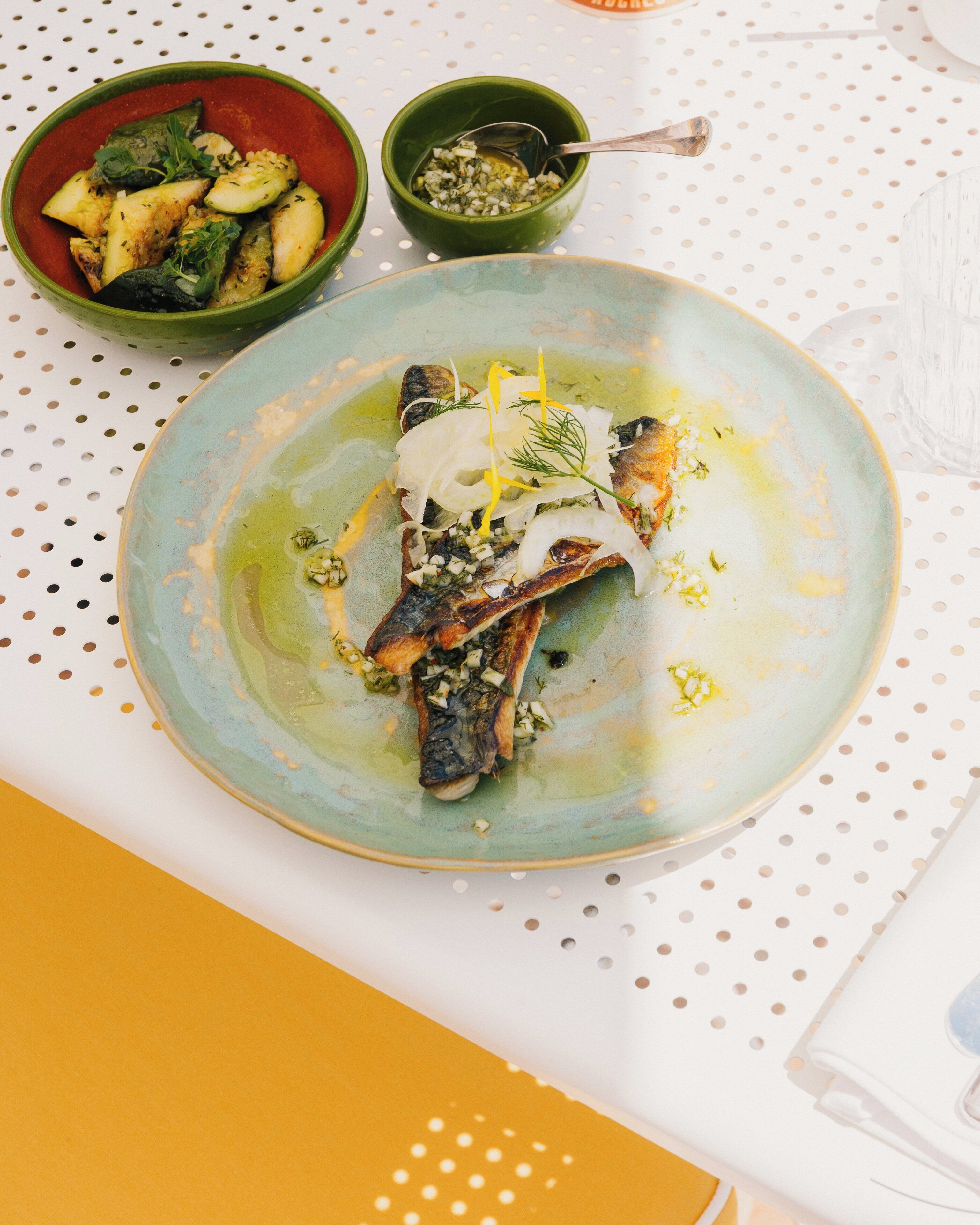 A plate of food with a green sauce, a bowl of vegetables, and a small bowl of sauce on a table with a white and yellow tablecloth.