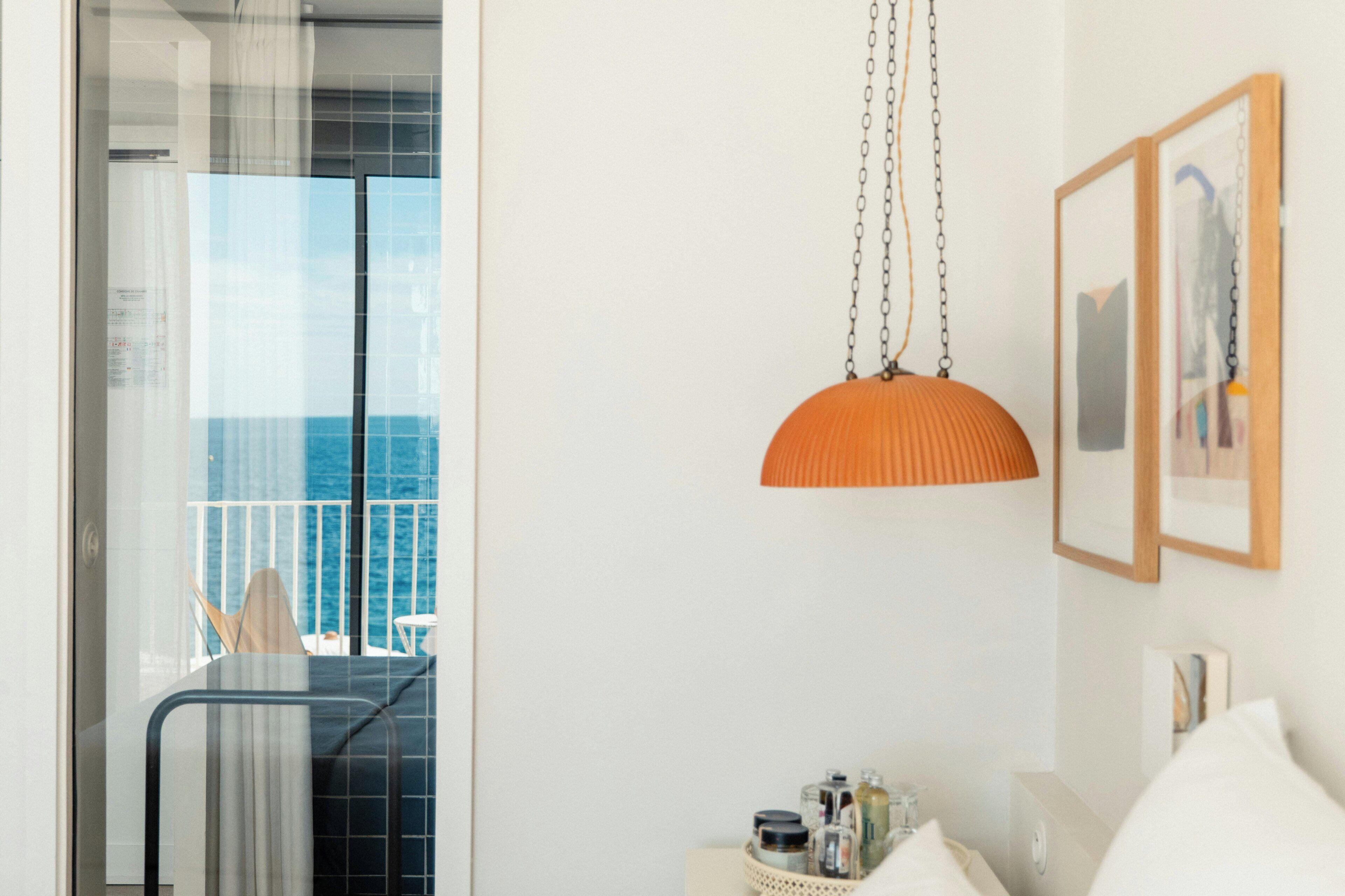 A hotel room with a balcony, a hanging orange pendant light, and framed artwork on the wall.