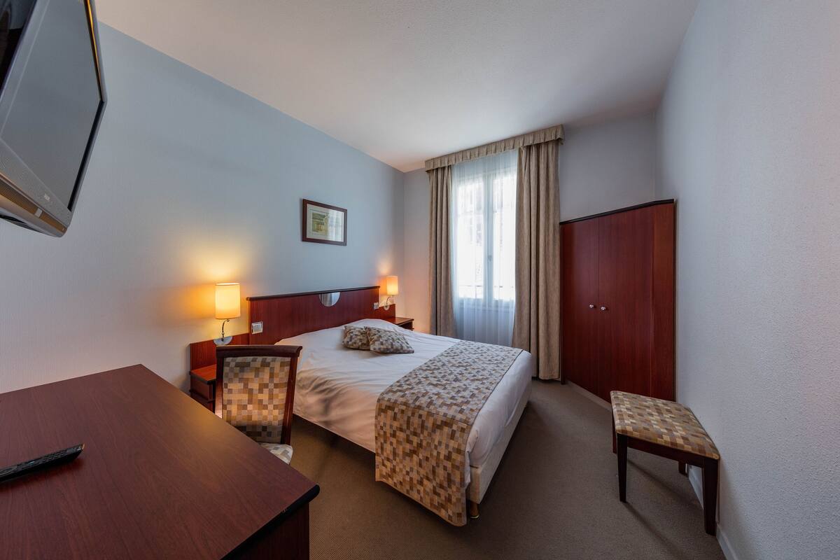 A hotel room with a bed, bedside tables, a desk, a chair, a wardrobe, and a television.