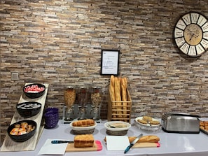 A buffet setup with various food items, including bread, pastries, and condiments, against a stone wall backdrop. - Noemys Néris Montluçon (Neris-les-Bains)