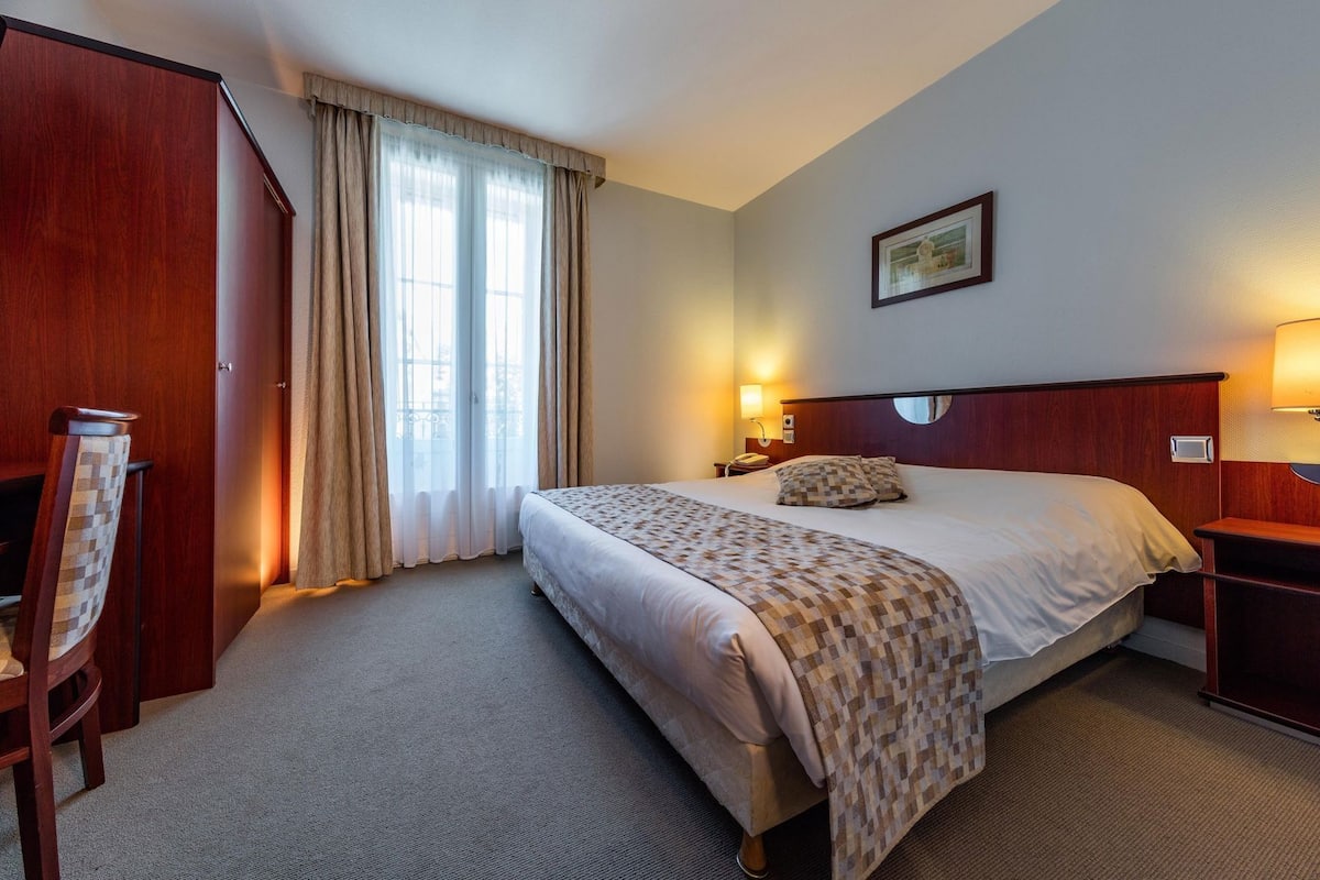 A hotel room with a bed, a desk, a chair, a nightstand, a lamp, and a window with curtains.