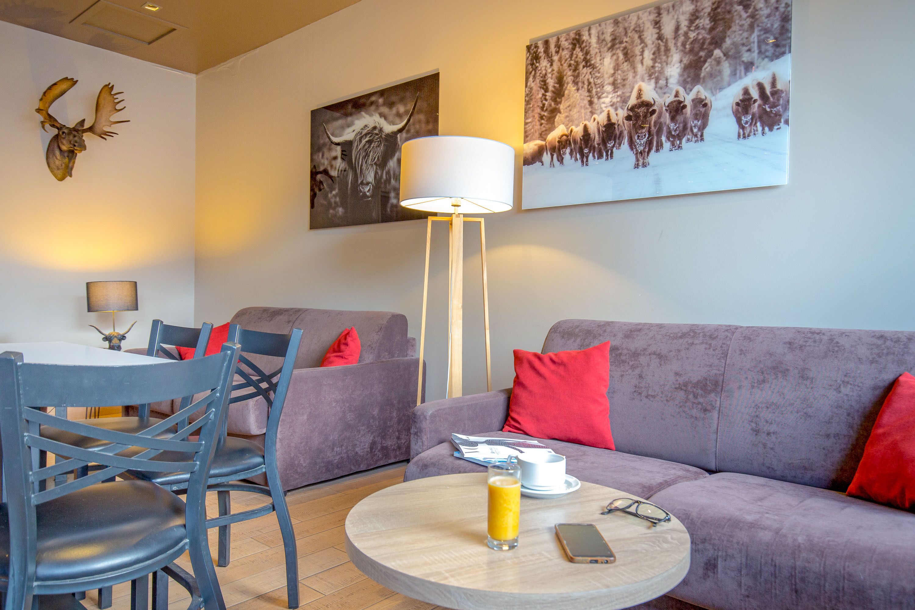 Un salon moderne avec un canapé gris, des coussins rouges, une table basse ronde sur laquelle se trouve un verre de jus de fruits, un téléphone portable et une lampe.