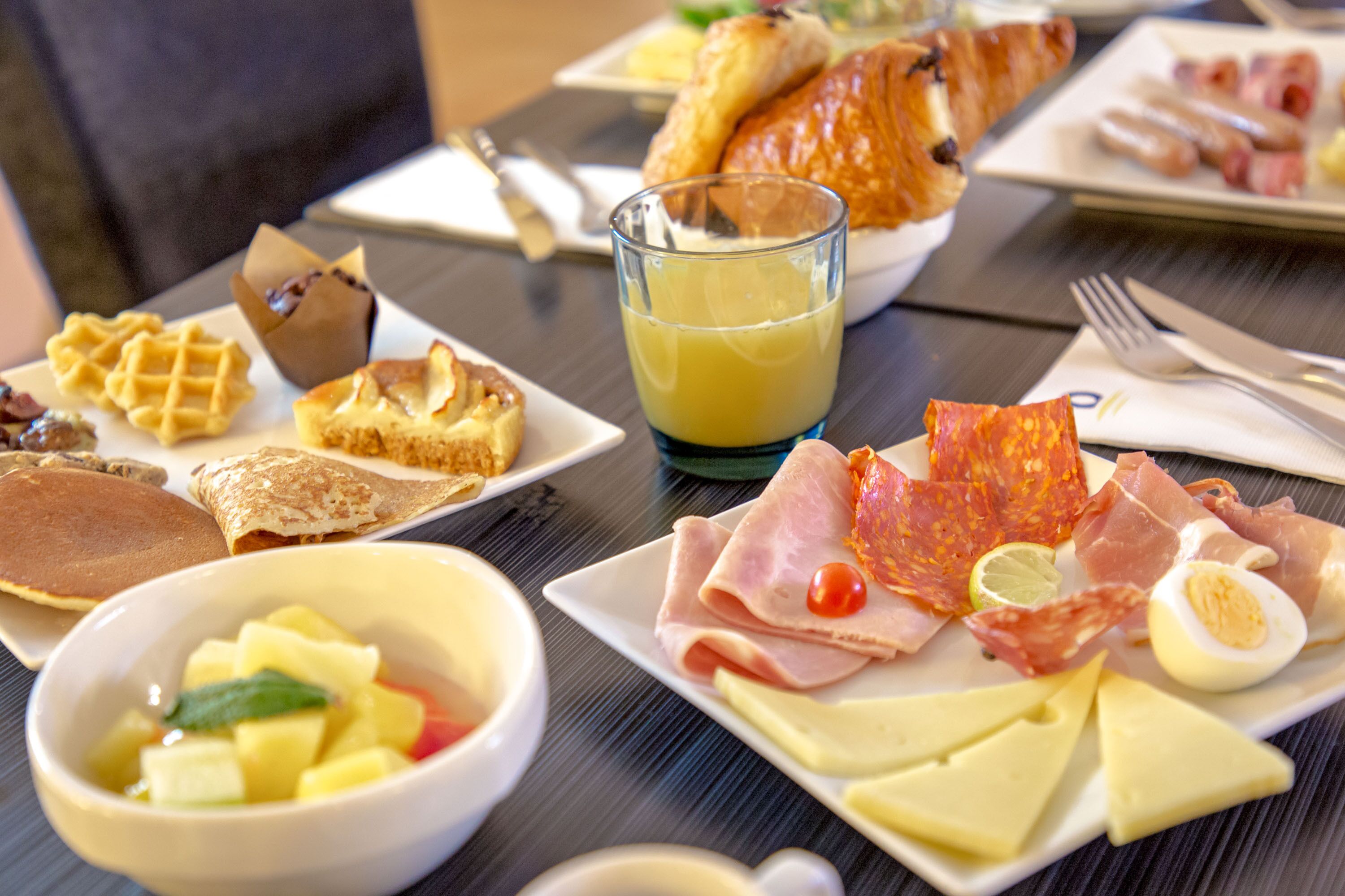 A breakfast table with waffles, pancakes, croissants, a glass of orange juice, and various meats and cheeses.