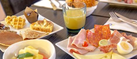 A breakfast table with waffles, pancakes, croissants, a glass of orange juice, and various meats and cheeses.