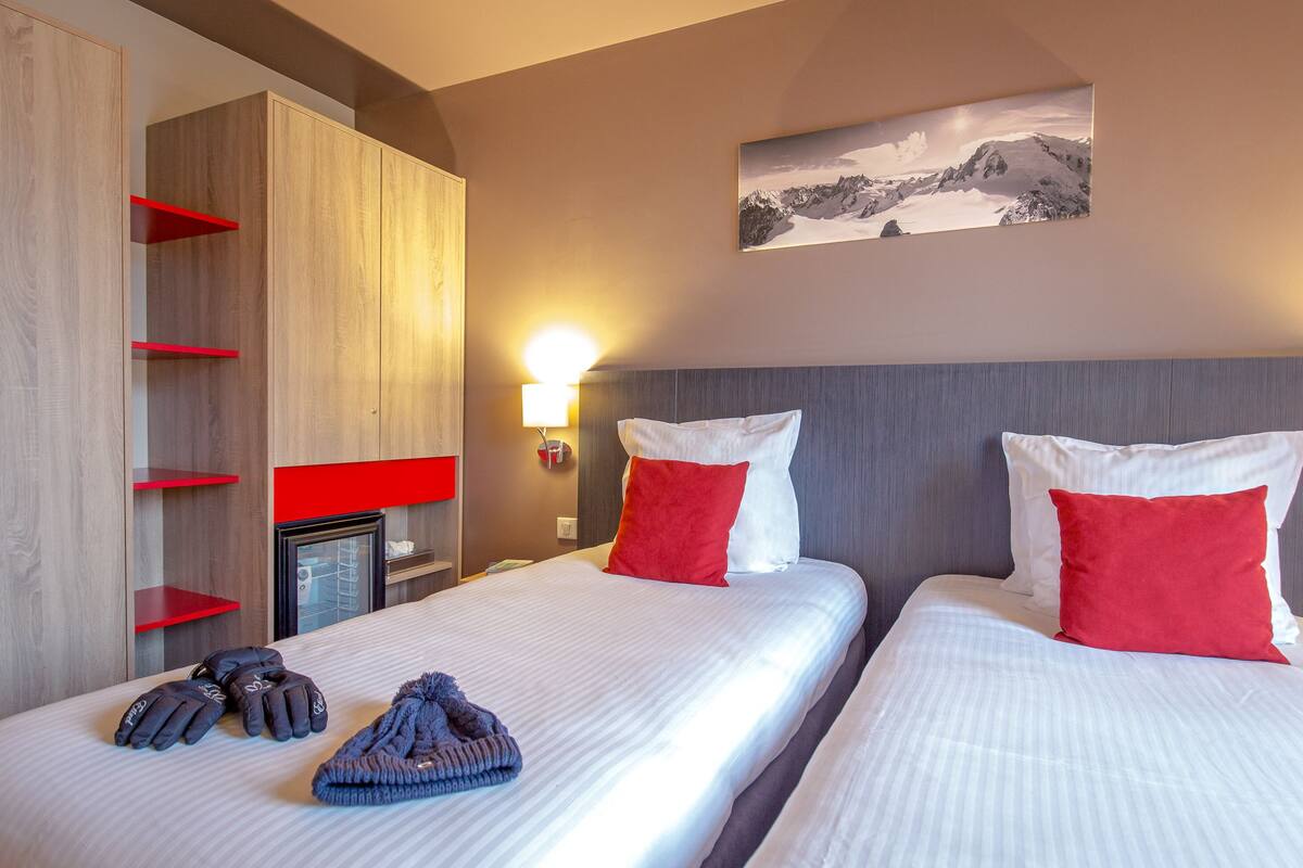 Ein Hotelzimmer mit zwei Betten, roten Kissen, einem Mantel und einem Gemälde einer Berglandschaft an der Wand.