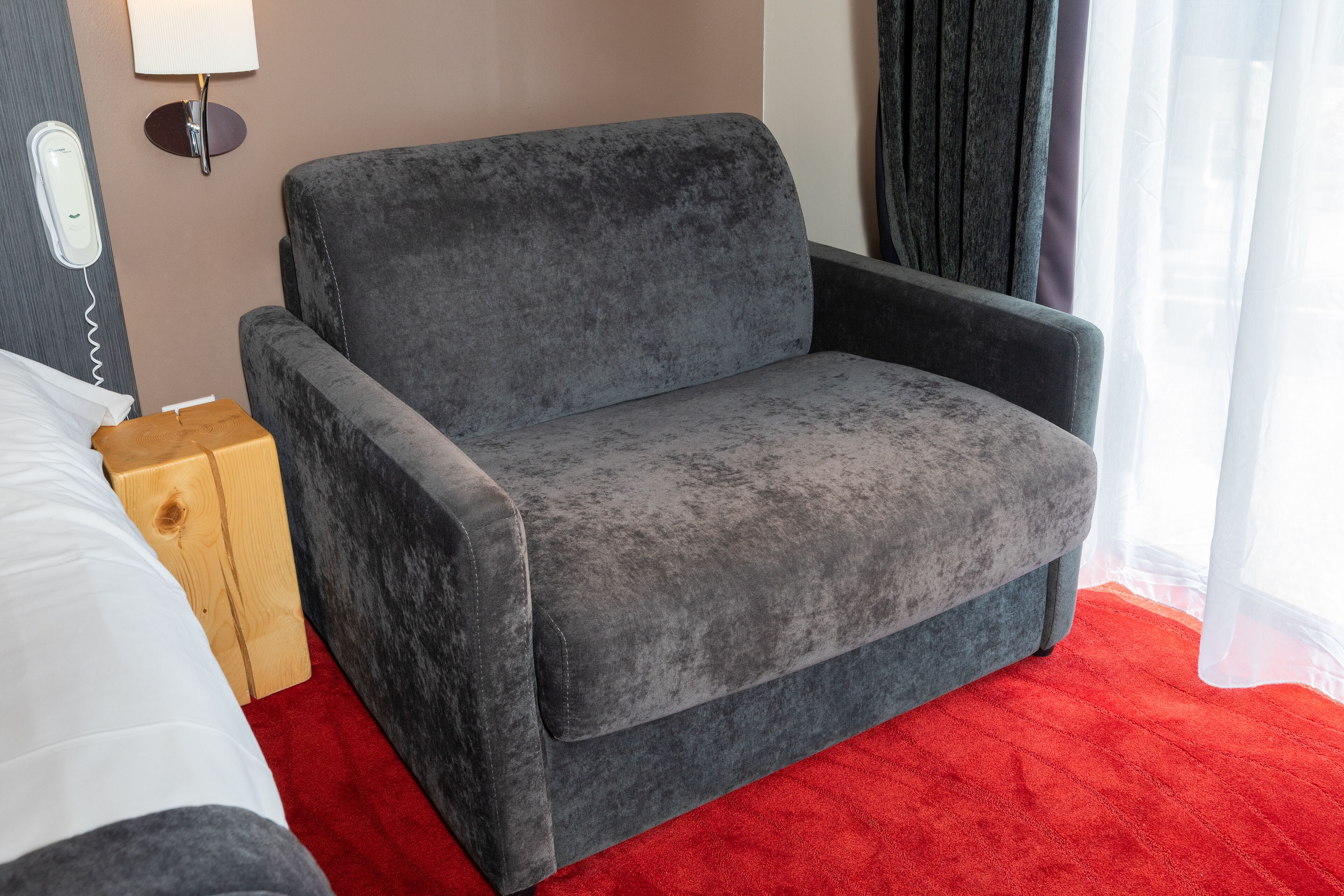 A grey sofa in a hotel room with a red carpet, a wooden nightstand, and a white bed.