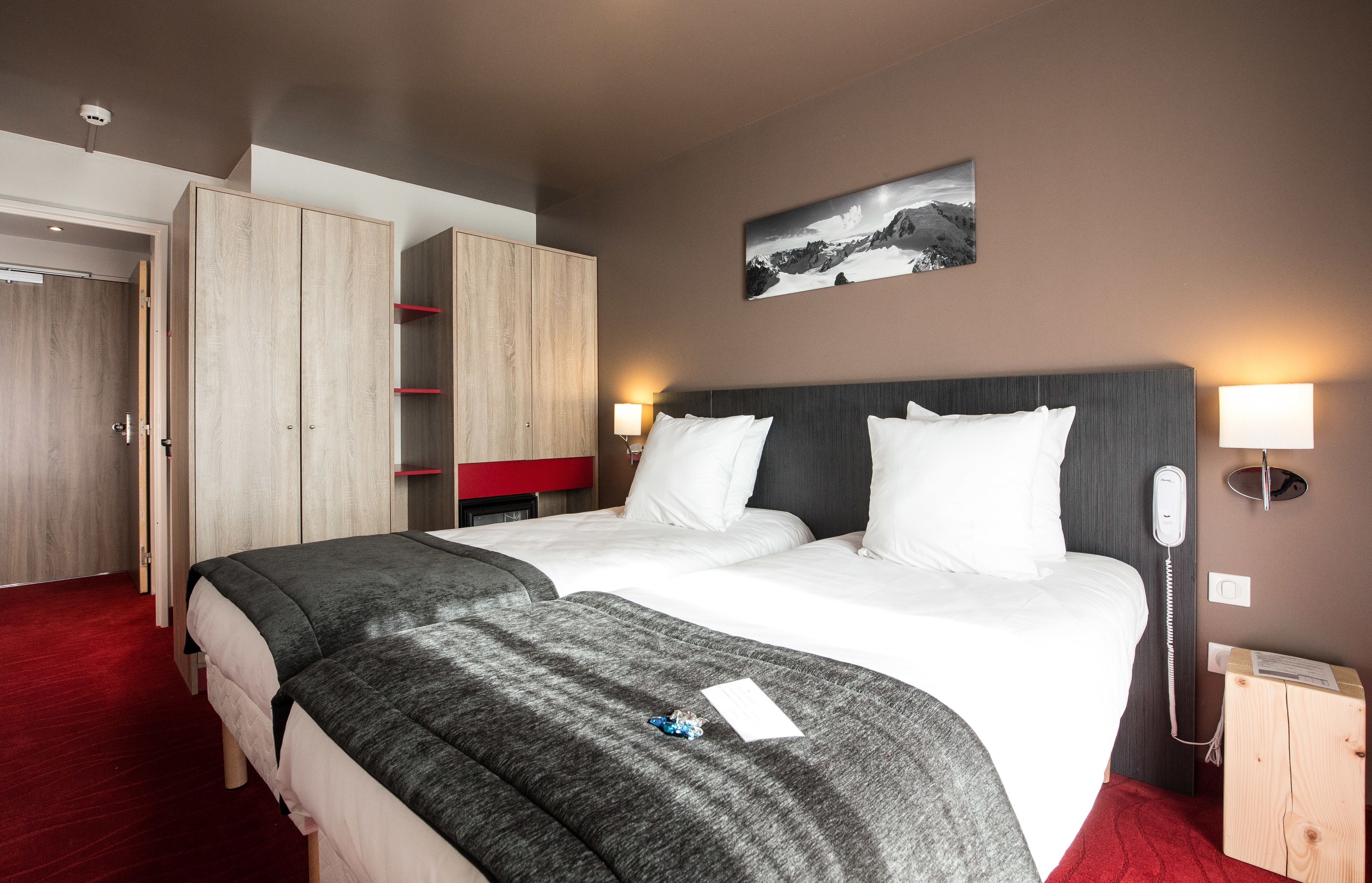 A hotel room with two beds, a wardrobe, a bedside table with a lamp, a phone, and a picture on the wall.