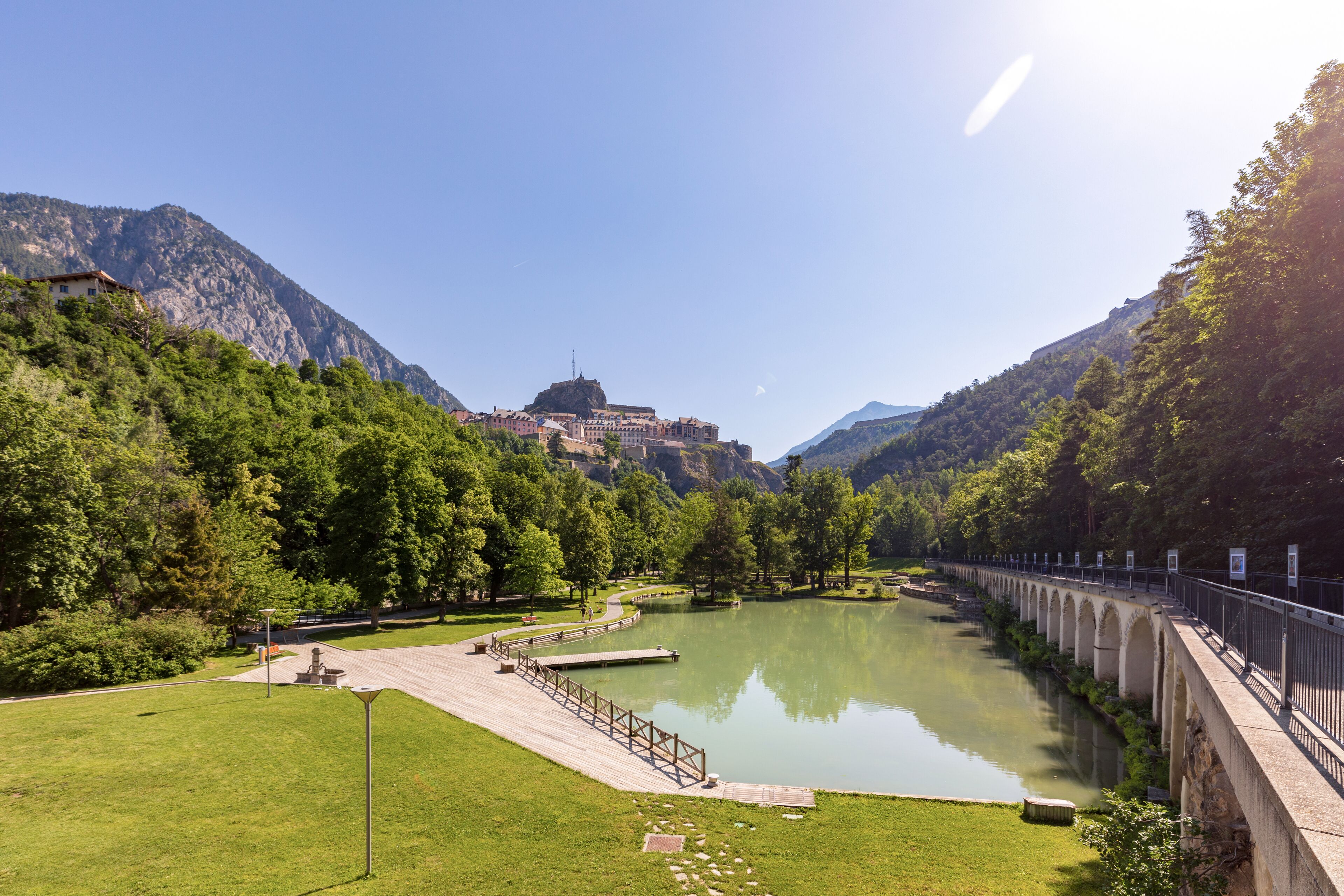 A serene riverside park with a bridge, lush greenery, and a mountain backdrop.