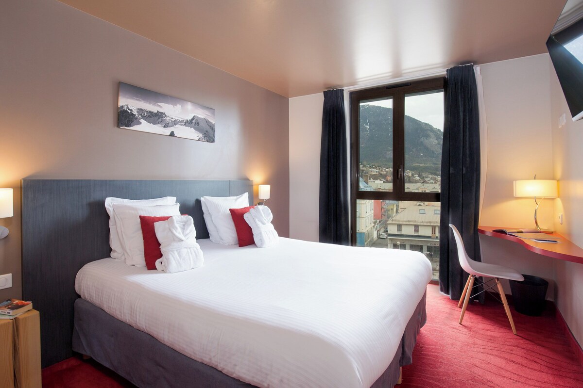 A hotel room with a large bed, two bedside lamps, a desk with a chair, a window with curtains, and a painting on the wall.