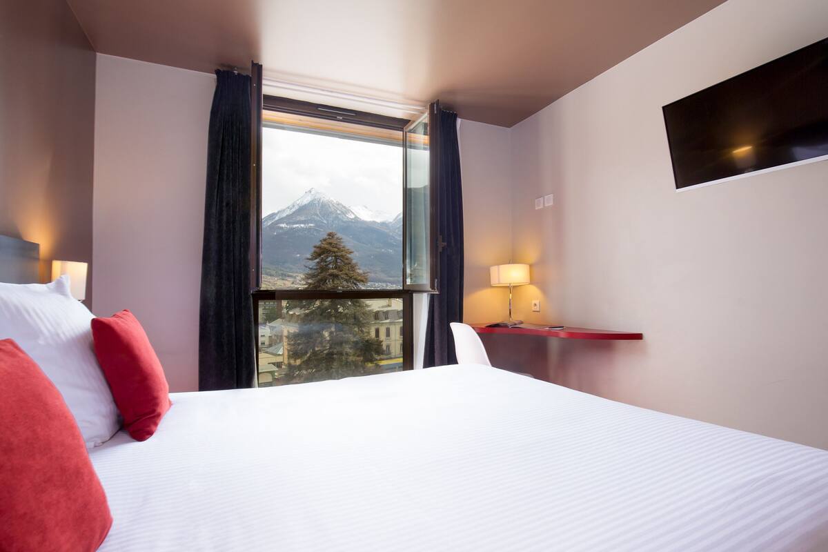 Ein Hotelzimmer mit einem großen Bett, einem Fenster mit Blick auf die Berge, einem Schreibtisch mit Lampe und einem an der Wand befestigten Fernseher.