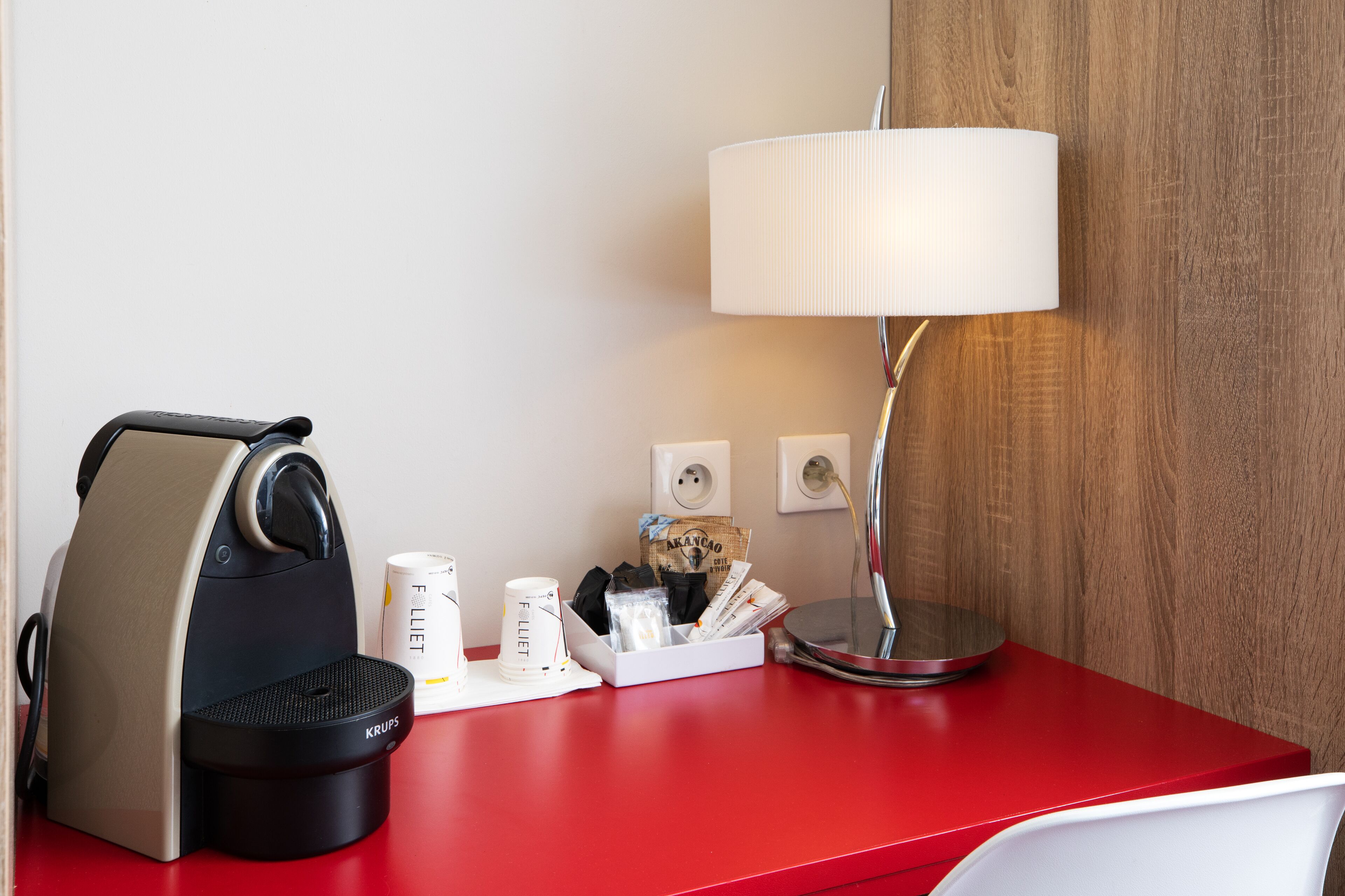 A red desk with a coffee maker, a lamp, and a cup holder with coffee cups and a bag of coffee.